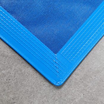 Nahaufnahme der Ecke eines Schutznetzes mit allseitig hochfrequent geschweißtem Saum oder Keder, blau und strukturiert, mit verstärkten Nähten auf einem grauen, glatten Betonboden.