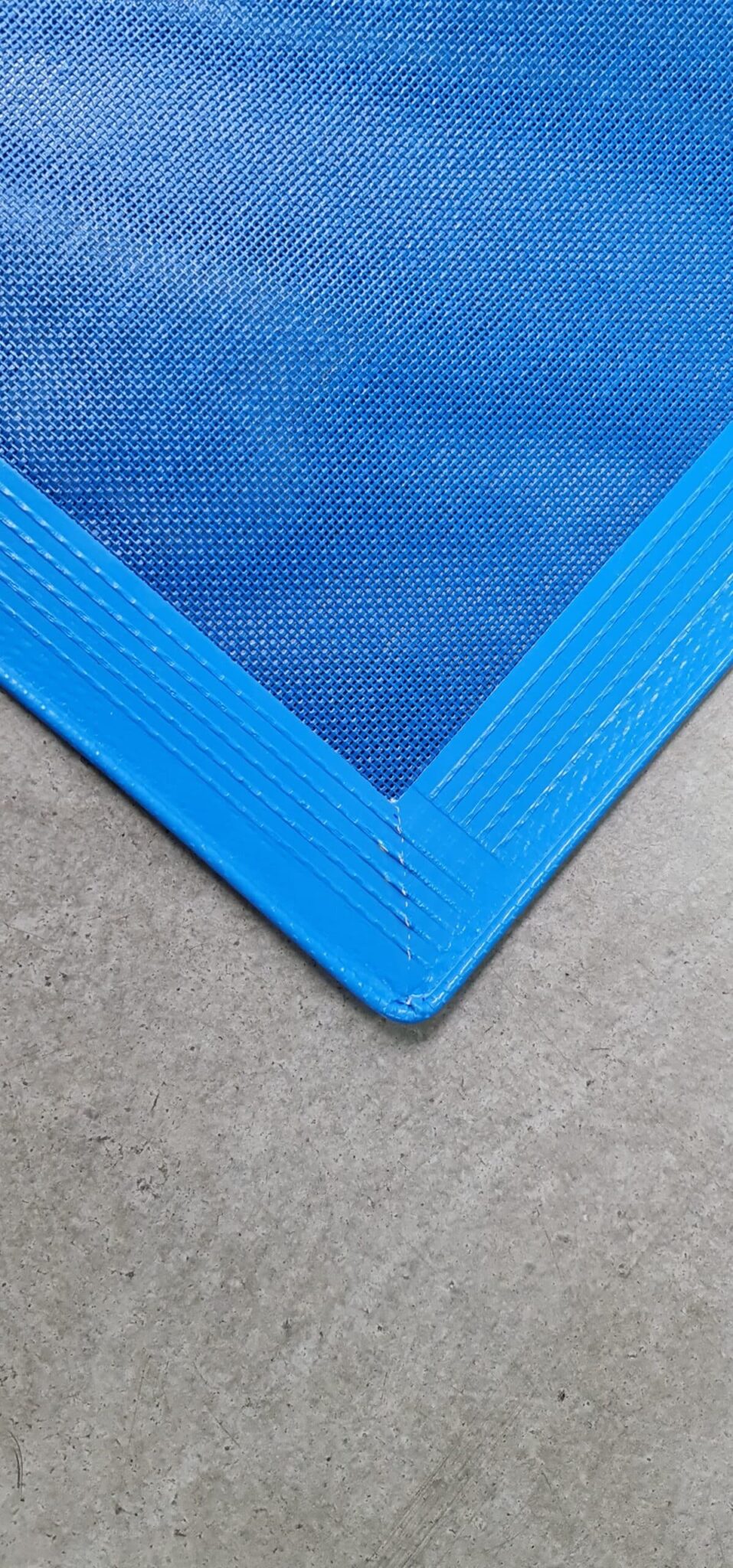 Nahaufnahme der Ecke eines Schutznetzes mit allseitig hochfrequent geschweißtem Saum oder Keder, blau und strukturiert, mit verstärkten Nähten auf einem grauen, glatten Betonboden.