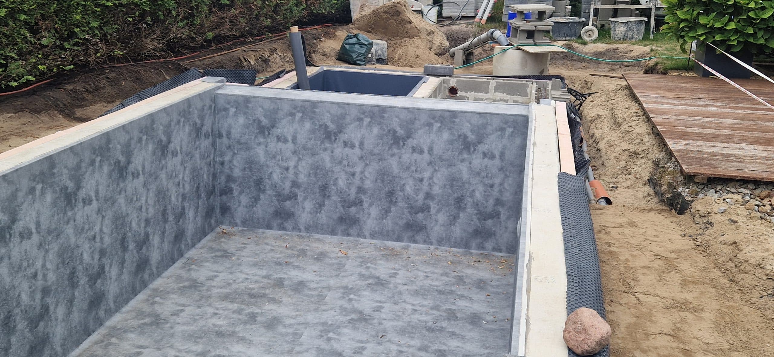 Nahaufnahme eines rechteckigen, unfertigen Schwimmbeckens mit grauem, strukturiertem Innenraum und verstreuten Baumaterialien. Das Schwimmbecken wird für eine Folienauskleidung vorbereitet. In der Nähe befinden sich Sand und Werkzeuge, rechts ist ein Holzsteg zu sehen.