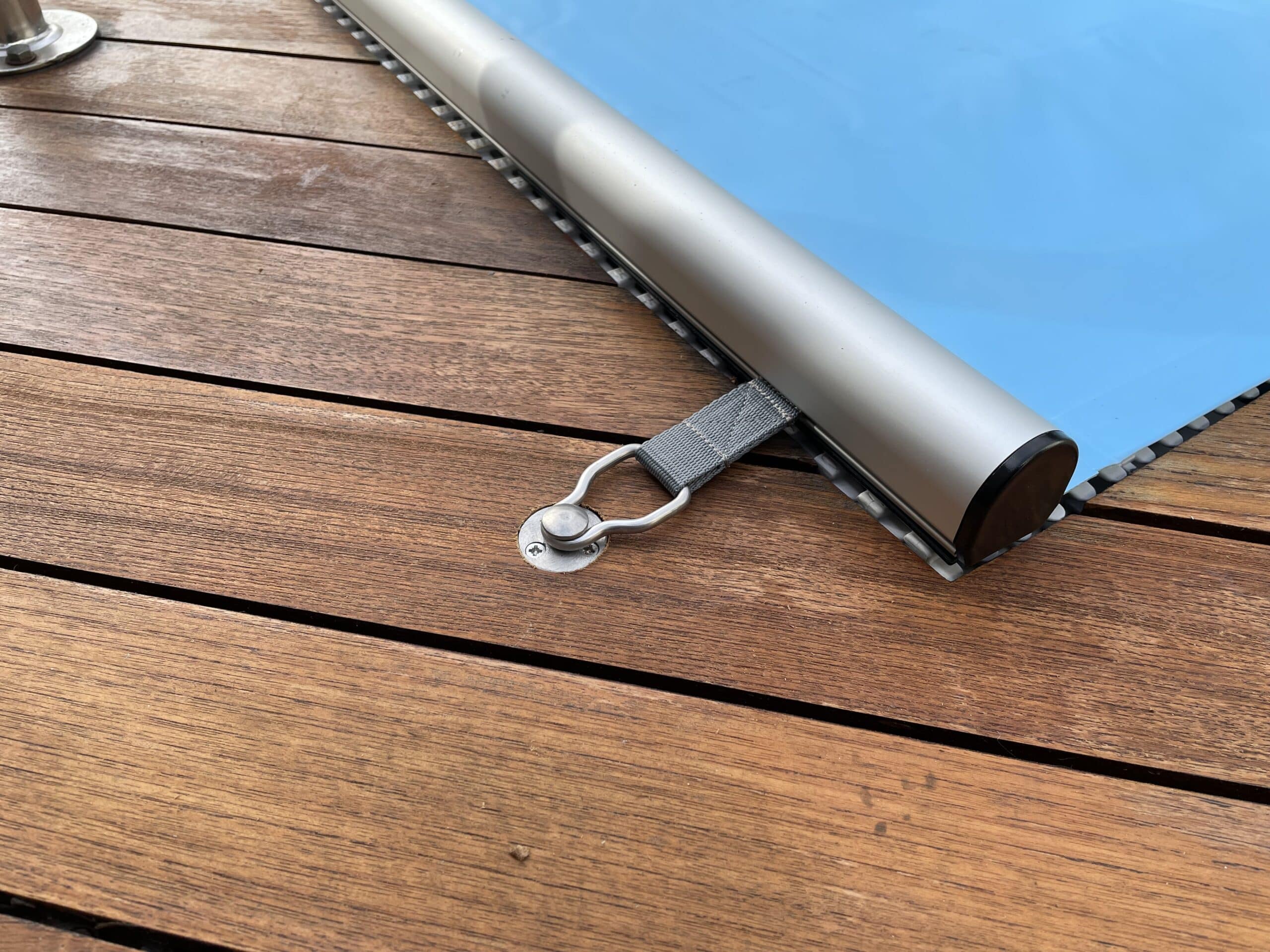 Nahaufnahme der Sicherheitsabdeckung Rollschutz Extra, einer blauen Schwimmbadabdeckung mit silbernem Metallrand und Verriegelung, die an einem Holzdeck befestigt ist; einschließlich Handkurbel und Befestigungsset für die Stirnseiten.