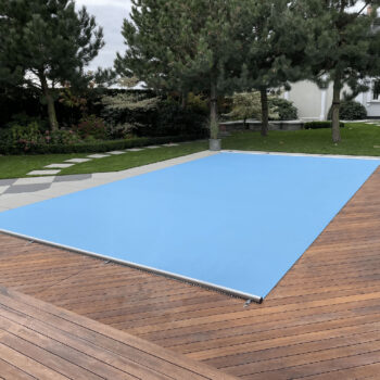 Ein rechteckiger Pool im Hinterhof ist mit der Rollschutz Extra Sicherheitsabdeckung inklusive Handkurbel und Befestigungs-Set für Stirnseiten gesichert, umgeben von einem Holzdeck, Bäumen und einem angelegten Garten. Ein Weg führt zu einem weißen Gebäude im Hintergrund.