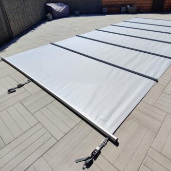 Eine Sicherheitsabdeckung Rollschutz inklusive Handkurbel und Befestigungs-Set für Stirnseiten in Weiß mit schwarzen Trägerstangen ist komplett über eine hellgraue Terrasse im Fischgrätmuster ausgebreitet. Im Hintergrund stehen Gartenmöbel und Pflanzkübel.