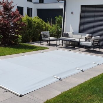 Eine Sicherheitsabdeckung / Rollschutz Standard inklusive Handkurbel und Befestigungs-Set für Stirnseiten schützt den kleinen eingelassenen Pool am modernen Haus, während nebenan Sitzmöbel auf der Terrasse stehen und der Garten das praktische Zubehör betont.