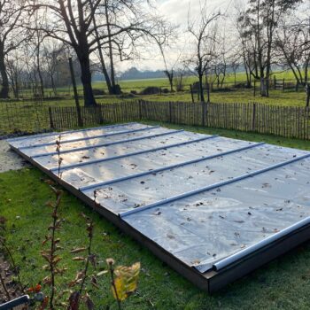 Ein rechteckiger Außenpool wird durch eine Sicherheitsabdeckung/Rollschutz inklusive Handkurbel und Befestigungs-Set für Stirnseiten geschützt. Gefallene Blätter liegen auf der Abdeckung, umgeben von Gras, blattlosen Bäumen und einem niedrigen Holzzaun.