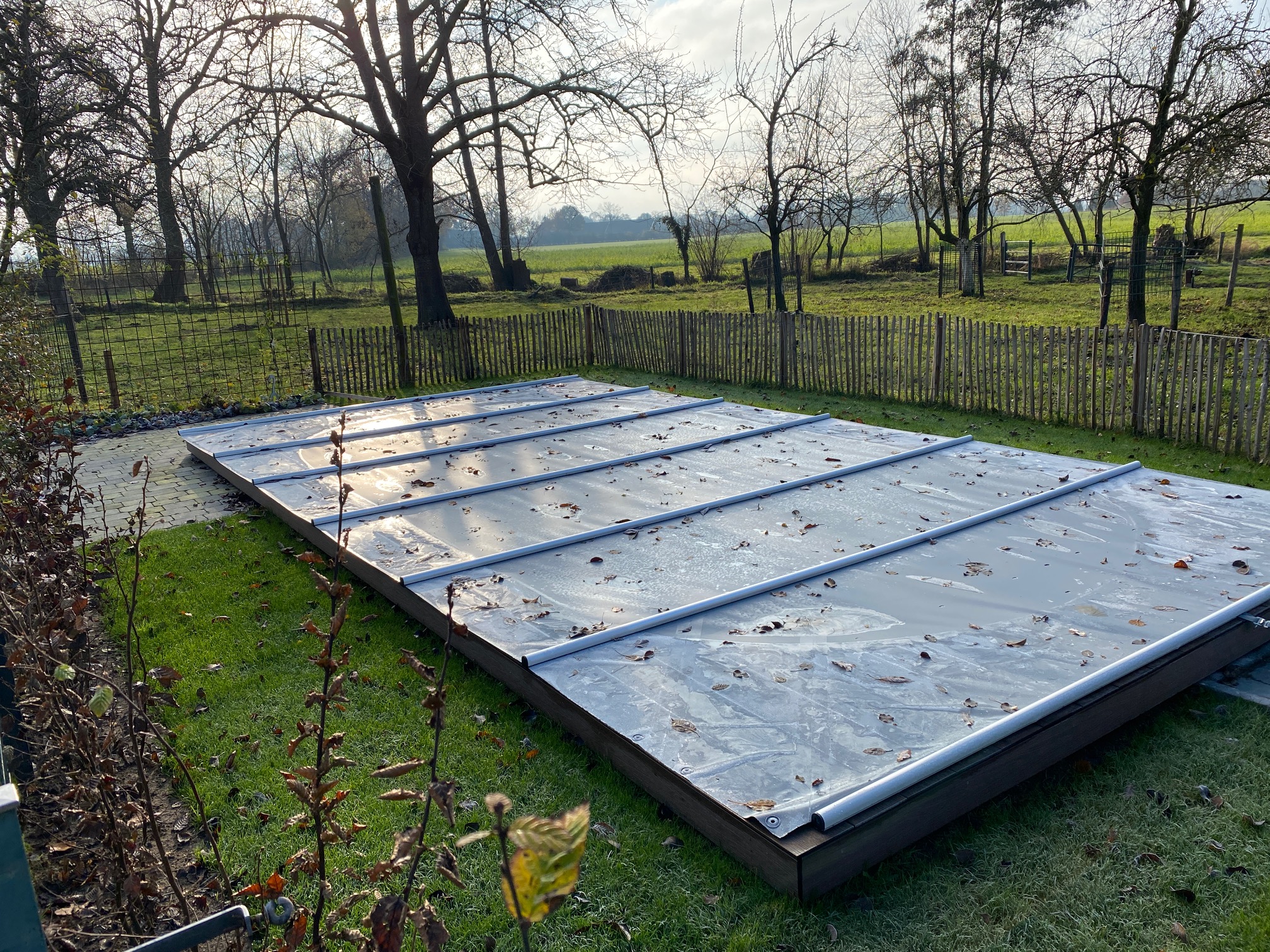 Ein rechteckiger Außenpool wird durch eine Sicherheitsabdeckung/Rollschutz inklusive Handkurbel und Befestigungs-Set für Stirnseiten geschützt. Gefallene Blätter liegen auf der Abdeckung, umgeben von Gras, blattlosen Bäumen und einem niedrigen Holzzaun.