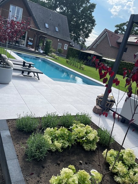 Ein moderner Garten mit einem mit hochwertiger Folie ausgekleideten Pool, einer gefliesten Terrasse, Liegestühlen, einem kleinen Gartenbeet mit Grünpflanzen und zwei Backsteinhäusern im Hintergrund an einem teilweise bewölkten Tag.