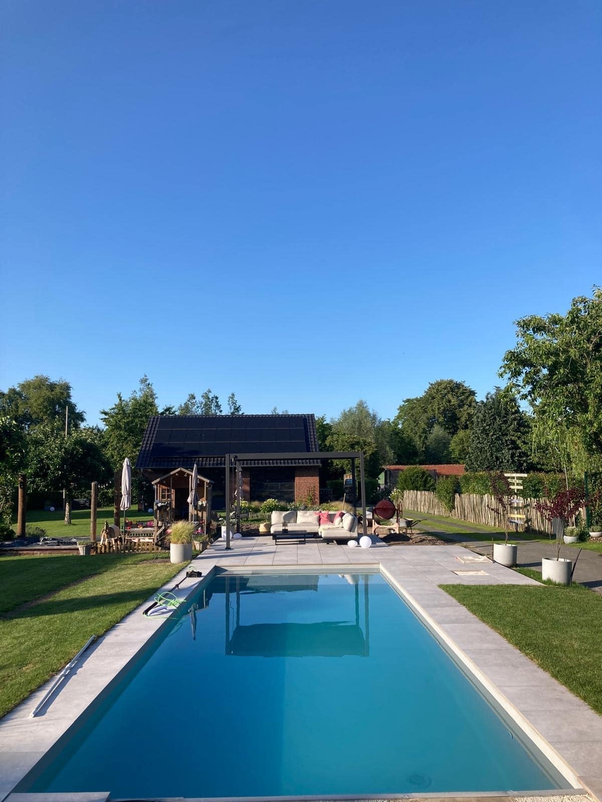Ein rechteckiger Außenpool mit einer eleganten Folienverkleidung ist von Gras und Topfpflanzen umgeben, mit einer modernen Terrasse und einem Sitzbereich im Hintergrund. Hohe Bäume und ein strahlend blauer Himmel vervollständigen die einladende Szenerie.