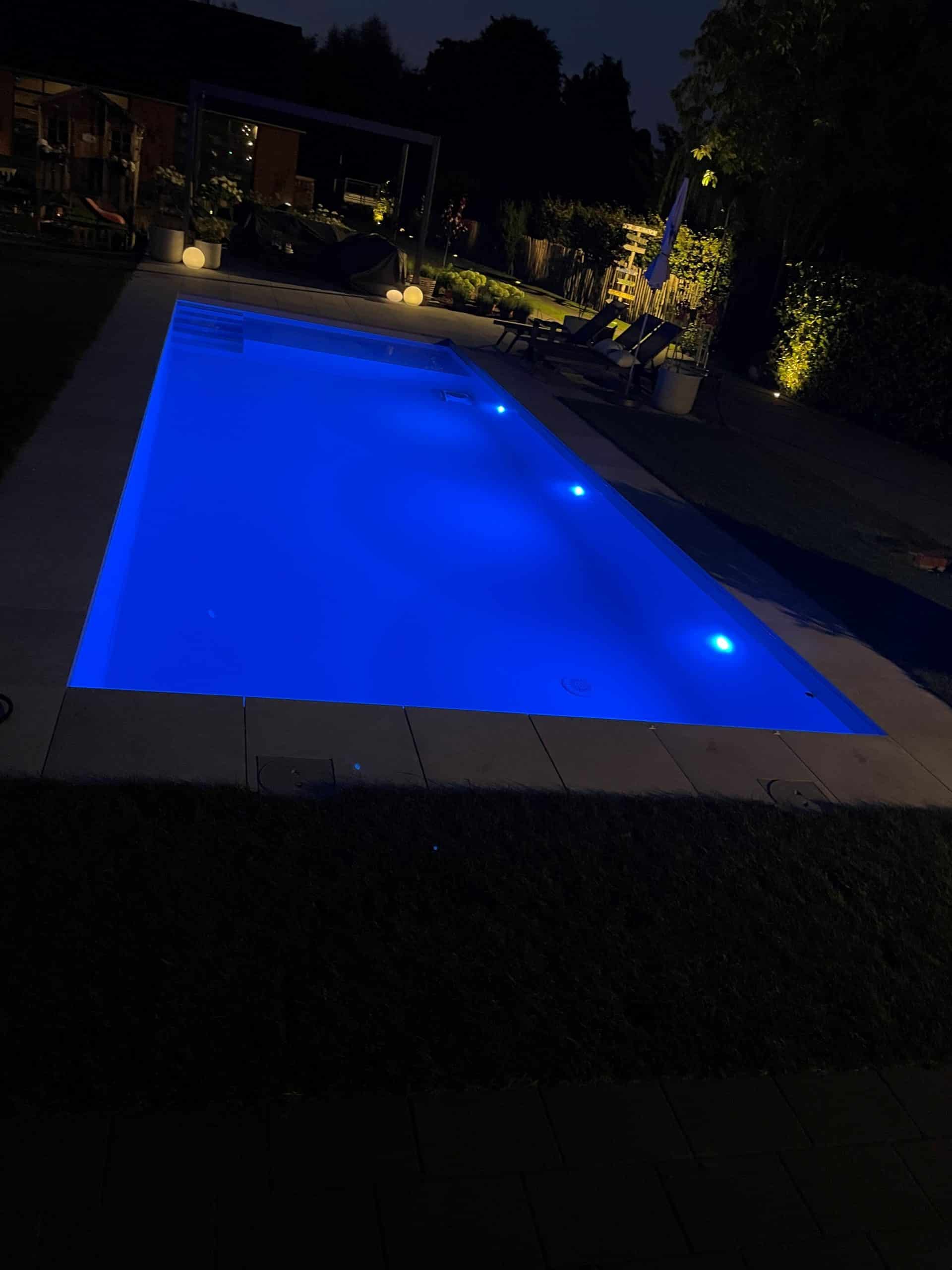 Ein rechteckiger Pool mit blauer Unterwasserbeleuchtung leuchtet in der Nacht. Die umliegende Terrasse und der Garten sind schummrig beleuchtet, mit Liegestühlen und Bäumen im Hintergrund. Das klare Wasser des Pools schimmert auf einer glatten Folie.
