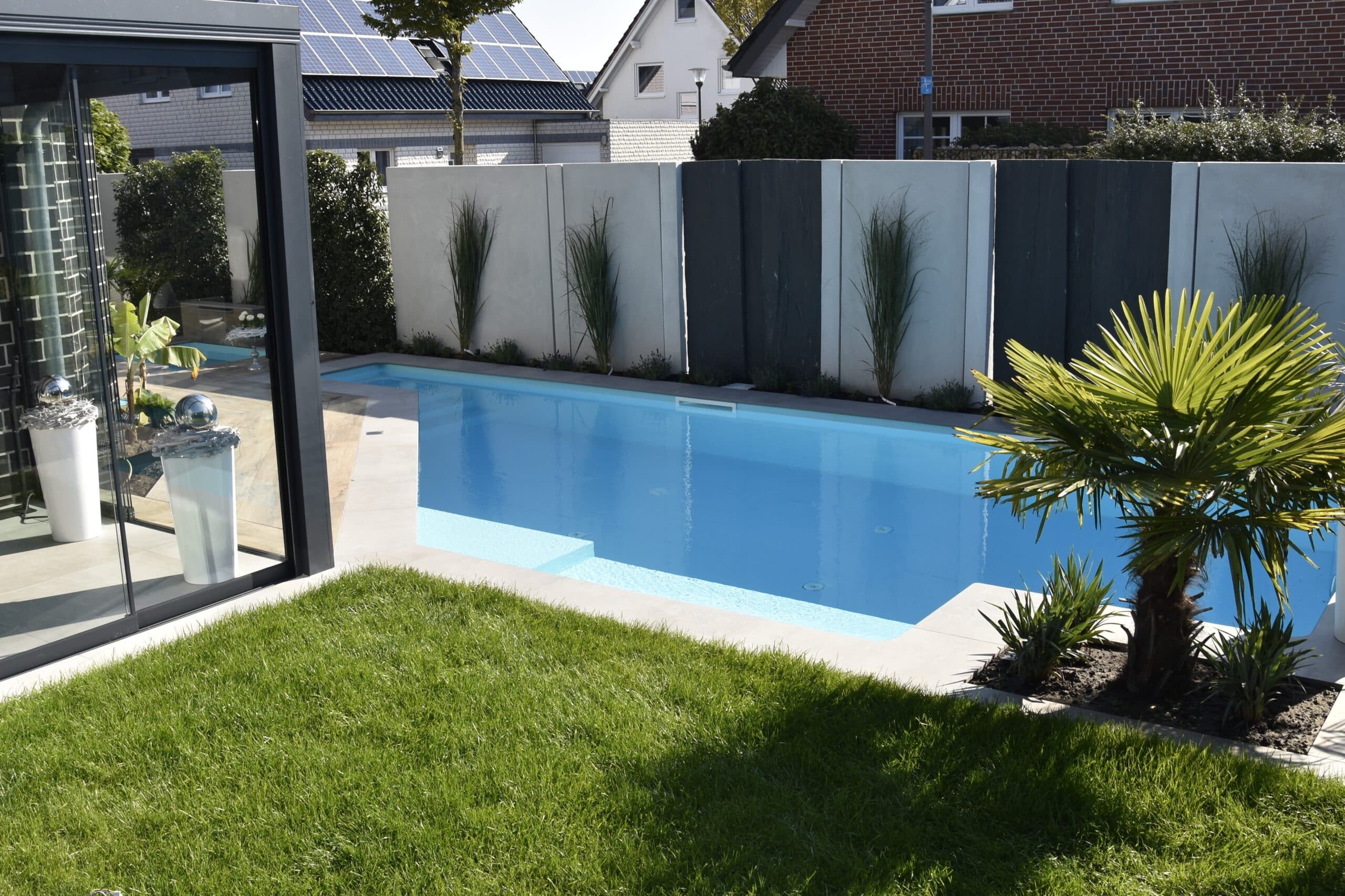 Ein kleiner rechteckiger Pool ist von Gras, ein paar Pflanzen und einem modernen Sichtschutzzaun umgeben. Auf der linken Seite sind ein Teil eines Hauses und eine Terrasse mit Glaswänden zu sehen.