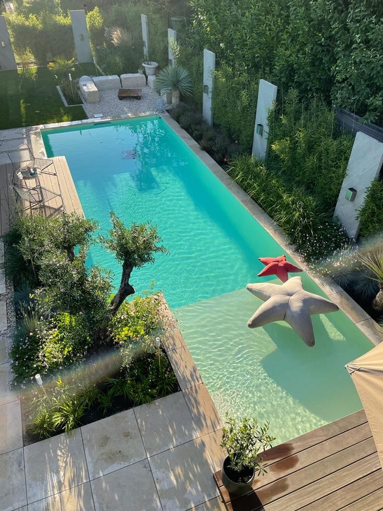 Ein rechteckiger Pool mit zwei großen sternförmigen Schwimmkörpern, umgeben von Grün und Steinfliesen, verfügt über ein Holzdeck im Vordergrund und stilvolle Gartenmöbel am anderen Ende.