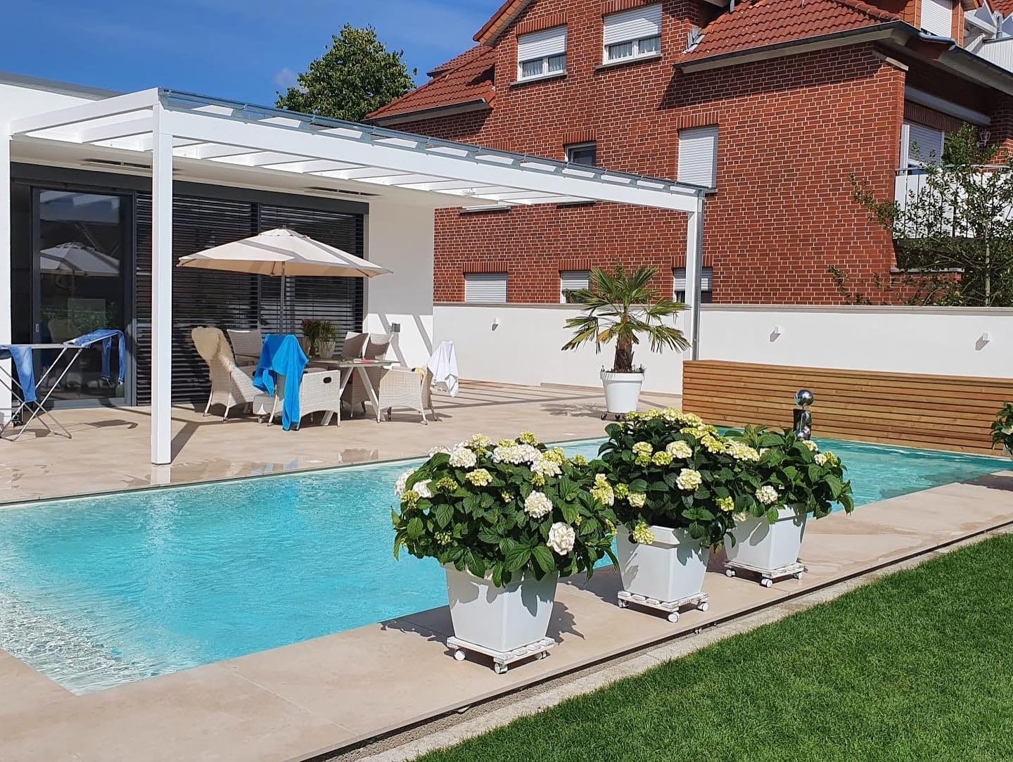 Ein moderner Hinterhof mit einem rechteckigen, mit Laubbäumen gesäumten Pool, drei getopften Hortensien auf der Terrasse, Liegestühlen mit Handtüchern, einem Sonnenschirm und einem roten Backsteinhaus im Hintergrund bei strahlend blauem Himmel.