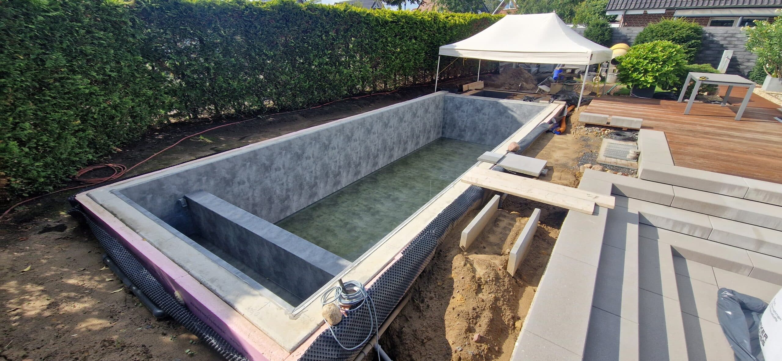 Rechteckiger, nicht fertiggestellter, mit Folie und Betonwänden ausgekleideter Pool, der teilweise mit Wasser gefüllt ist. Auf dem Gelände liegen Baumaterialien, eine Holzterrasse, ein weißes Vordach und dichte grüne Hecken.