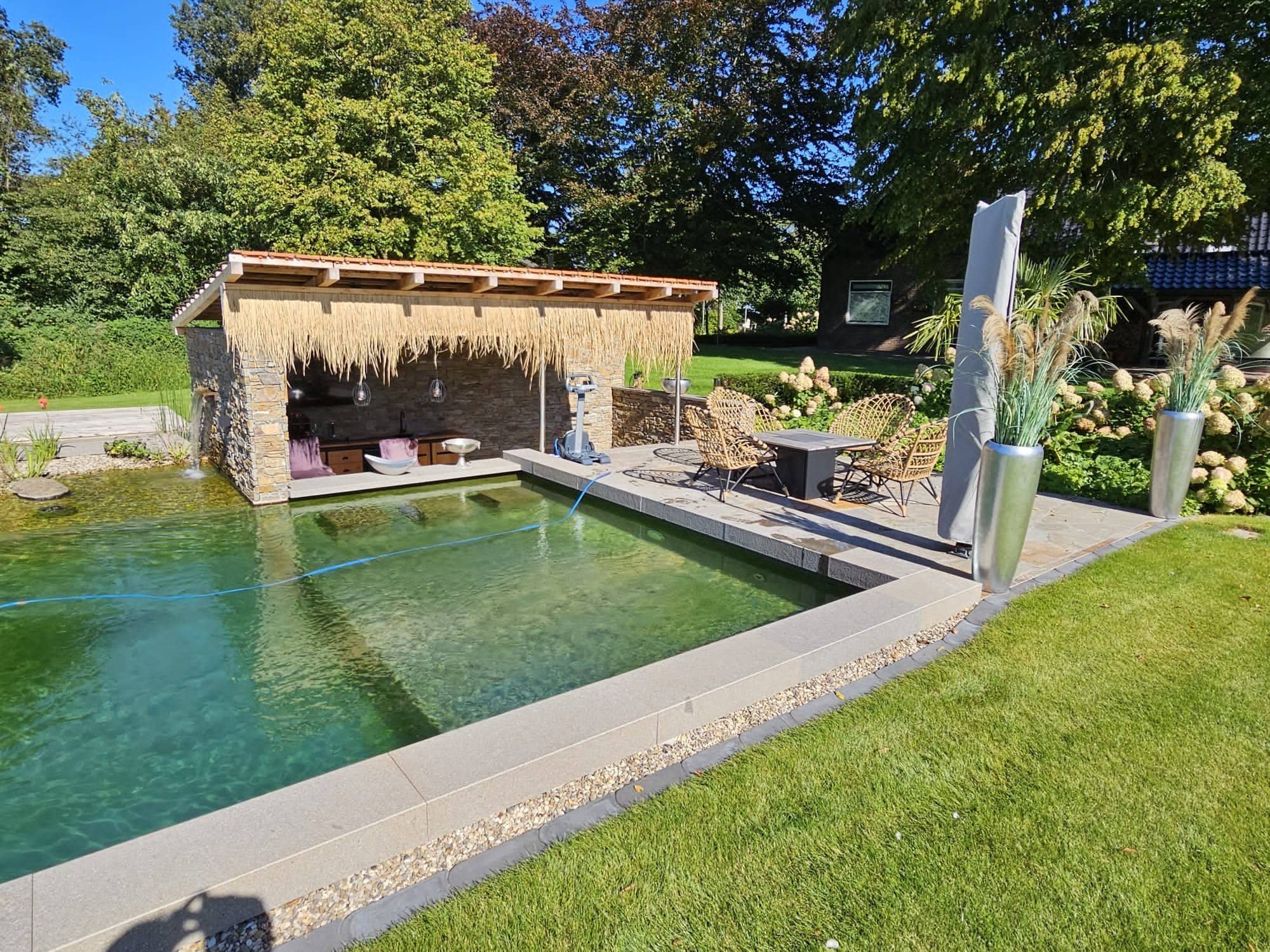 Ein moderner Garten mit einem rechteckigen Pool, der mit hochwertiger Folie ausgekleidet ist, einem überdachten Lounge-Bereich mit Korbmöbeln, einer Steinmauer, einem Esstisch für vier Personen auf der Terrasse und großen Pflanzgefäßen mit Ziergräsern.