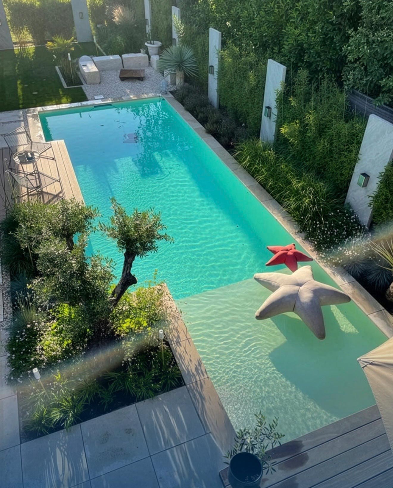 Ein rechteckiger Gartenpool mit klarem, blauem Wasser, umgeben von Grünpflanzen und Sitzgelegenheiten. Zwei große sternförmige Schwimmkörper, ein roter und ein grauer, befinden sich im flachen Ende. Der Bereich ist sonnig und ordentlich bepflanzt.