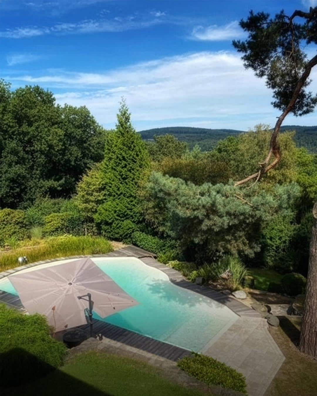 Ein Pool im Hinterhof, umgeben von Grün, mit einem großen Sonnenschirm, der das Pooldeck teilweise beschattet. Bäume und dichtes Blattwerk umschließen das Gebiet, und in der Ferne sind Hügel unter einem blauen Himmel mit vereinzelten Wolken zu sehen.