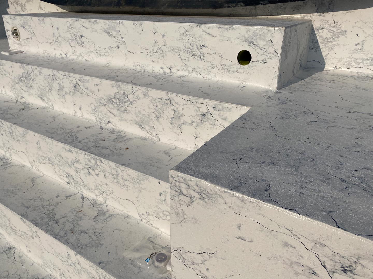 Weiße Marmorstufen mit grauer Maserung, mit einem kleinen kreisförmigen Loch auf der vertikalen Fläche einer oberen Stufe; auf den unteren Stufen sind eine weggeworfene Plastikflasche und ein Deckel zu sehen.