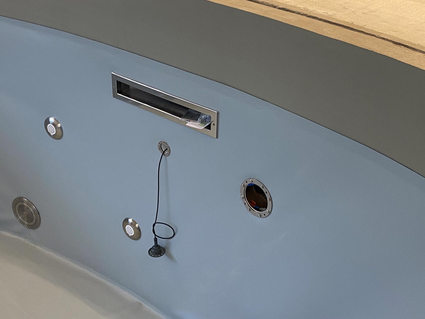 Nahaufnahme der Seitenwand einer Badewanne mit einem Kabel, das von einem Stecker herabhängt, drei runden Bedienknöpfen, einem rechteckigen Metallschlitz und einer offenen runden Öffnung. Das Wanneninnere und der Wannenrand sind sichtbar.