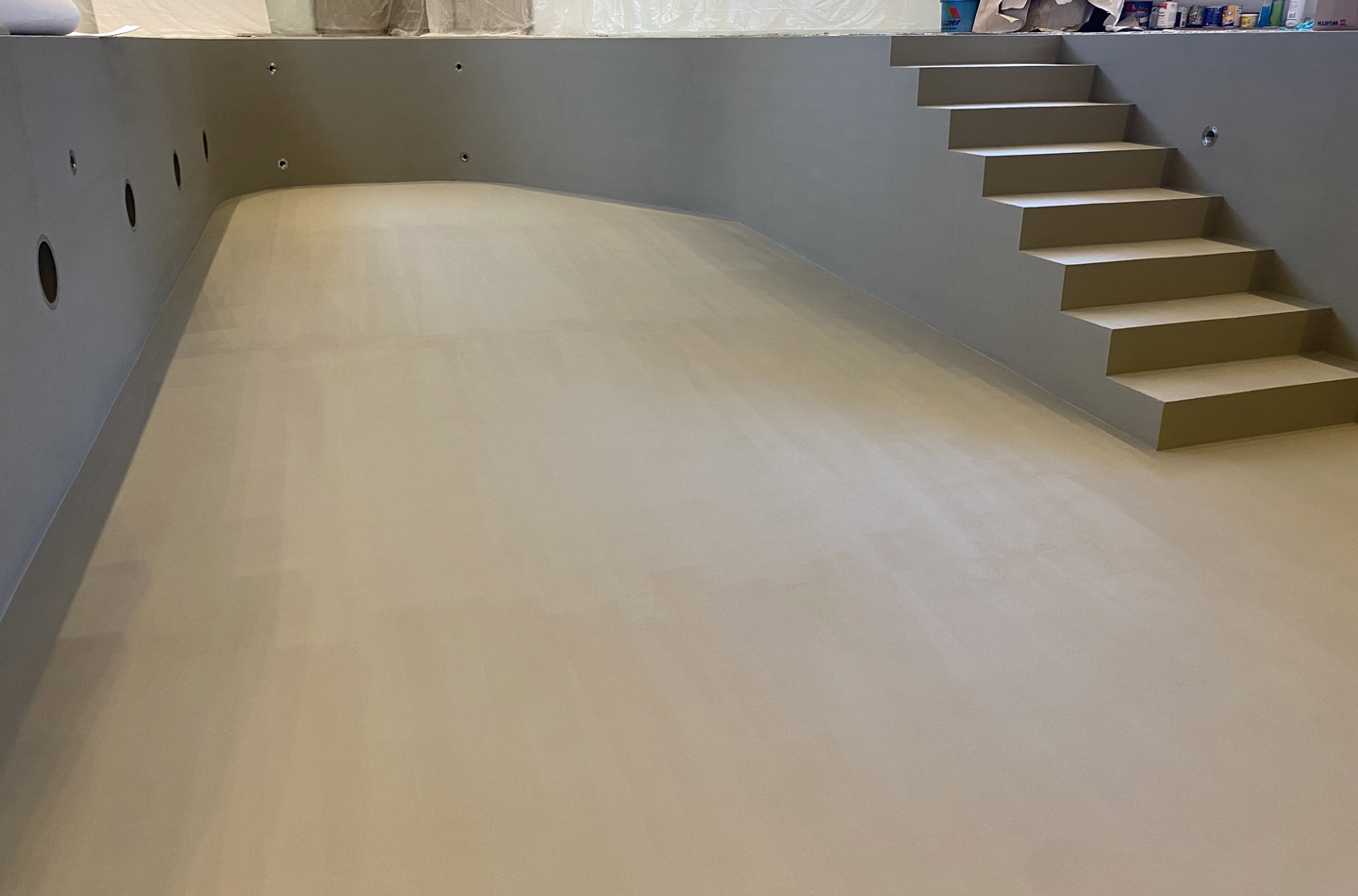 Ein leeres Hallenbad mit glatten, beigen Wänden und Böden, mehreren kreisförmigen Abflussöffnungen an einer Seite und einer Reihe von rechteckigen Stufen, die in das Becken führen. Auf der Oberfläche oberhalb der Stufen sind Vorräte sichtbar.