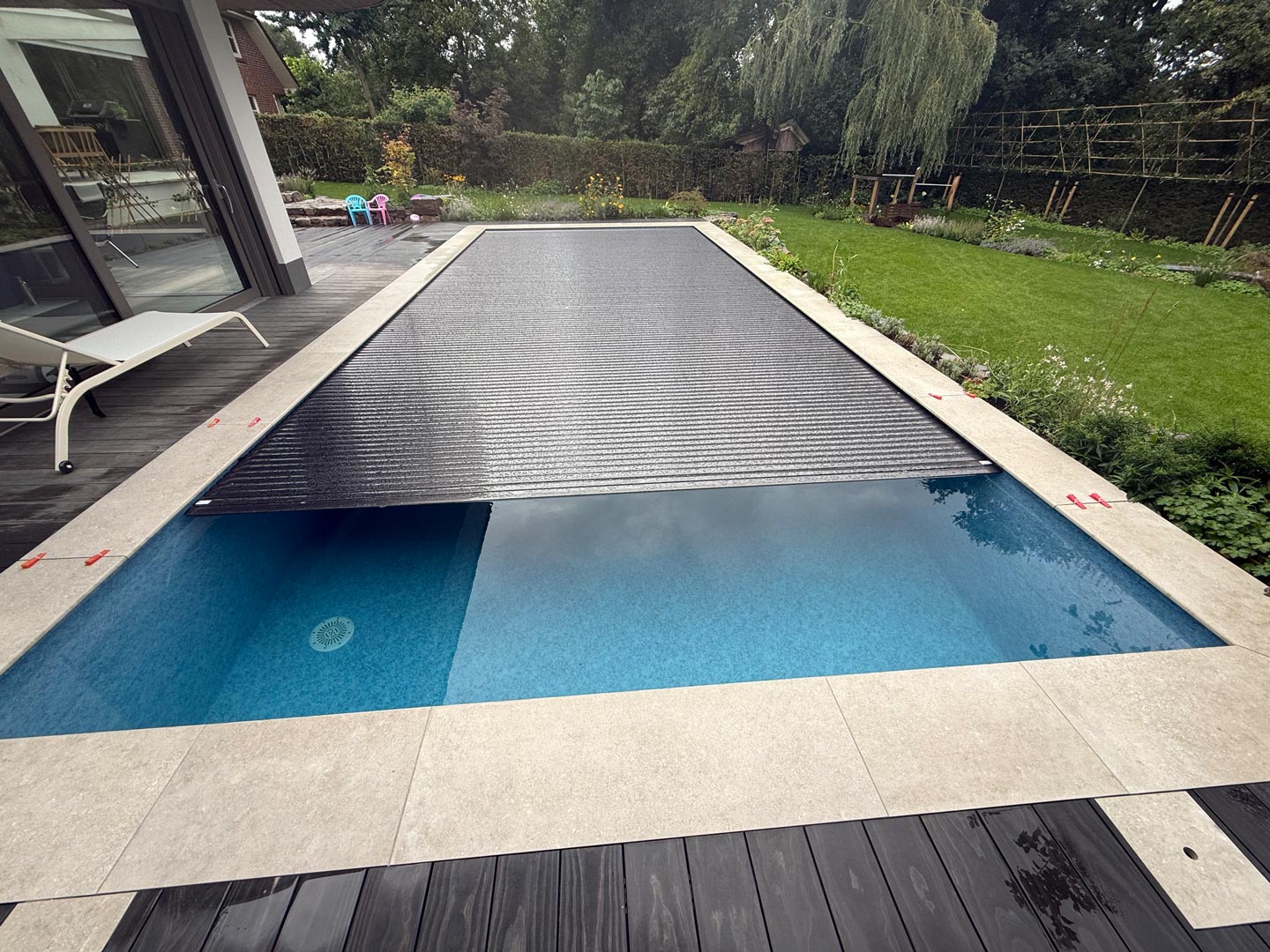 Der rechteckige Pool im Hinterhof ist teilweise von einer schwarzen, mit Lamellen versehenen Poolabdeckung bedeckt. Der Pool ist von hellen Steinfliesen umgeben und grenzt an eine Terrasse und einen grasbewachsenen Gartenbereich mit Pflanzen und Bäumen.