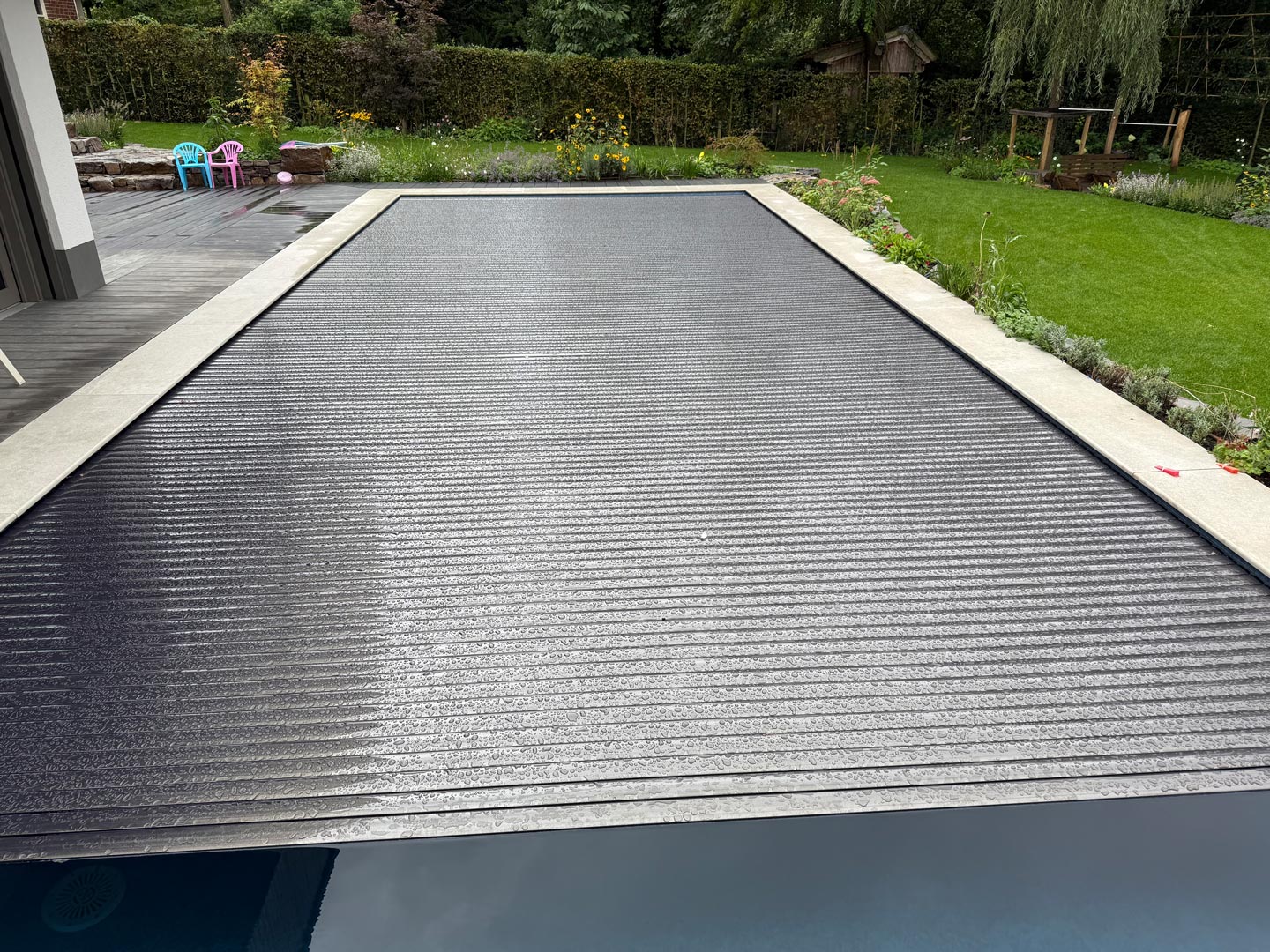 Ein rechteckiges Schwimmbecken ist mit einer dunklen, geriffelten Sicherheitsabdeckung versehen. Der Pool ist von einer Betonumrandung, einer Holzterrasse, Rasen und einem Garten umgeben, in dem im Hintergrund Bäume und Outdoor-Spielzeug zu sehen sind.