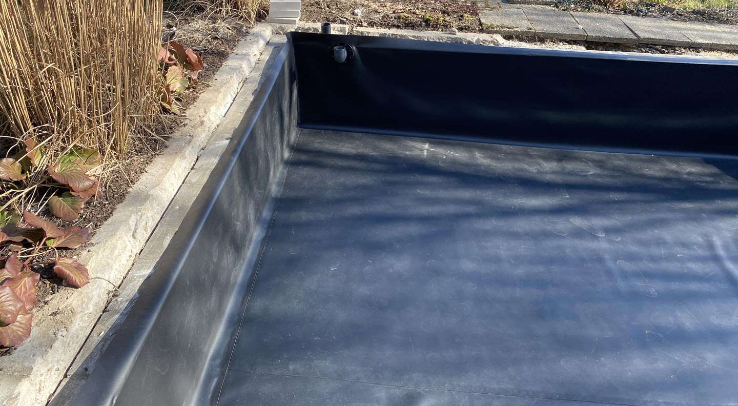 Ein rechteckiger, leerer Teich oder ein Wasserspiel, das mit schwarzem Material ausgekleidet ist, befindet sich neben einer Steinumrandung, trockenen Pflanzen und Pflastersteinen in einer Gartenanlage.