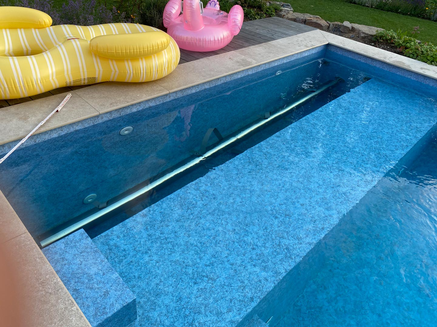 Ein Swimmingpool mit klarem, blauem Wasser, eine gelb gestreifte, aufblasbare Liege und ein rosa Flamingo schwimmen auf dem Pooldeck. Im Hintergrund befindet sich ein Garten mit Gras und Blumen.