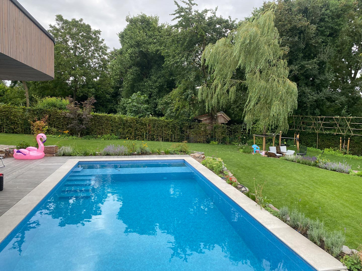Ein rechteckiger Außenpool mit klarem, blauem Wasser ist von einem Garten mit grünem Gras, Bäumen und Pflanzen umgeben. Ein rosa Flamingo-Poolschwimmer steht am Beckenrand, und zwei Liegestühle stehen auf dem Rasen.