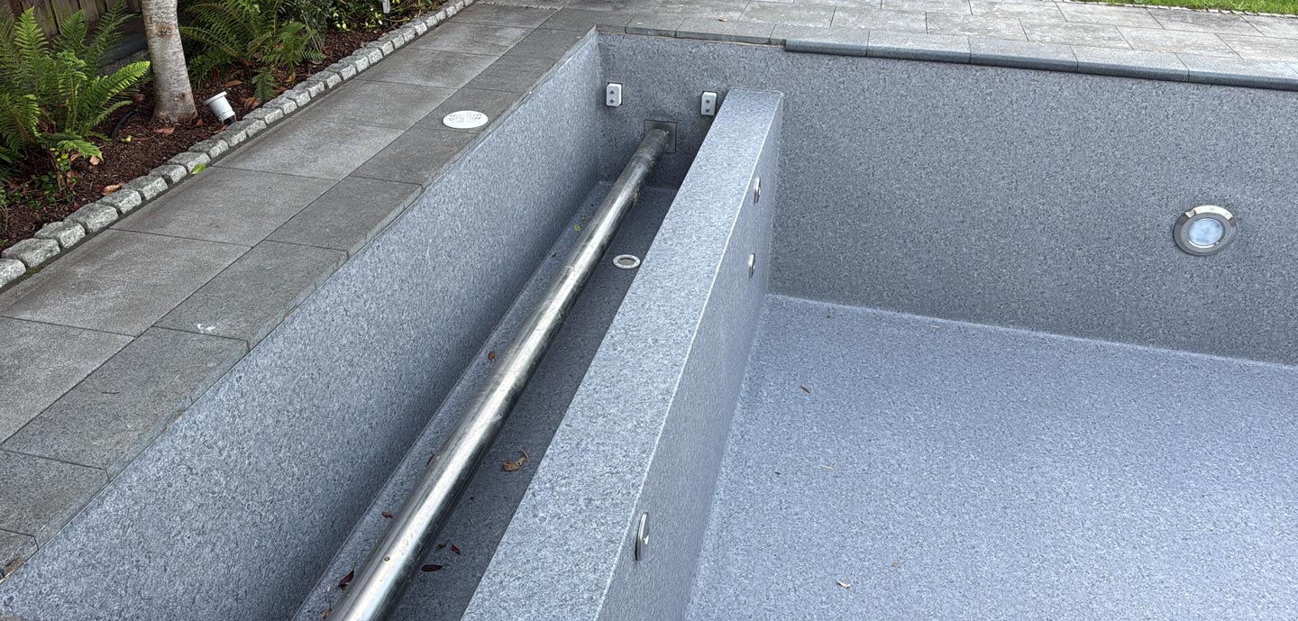 Ein leeres, graues Schwimmbecken mit einer eingebauten Trennbank und einem Metallgeländer. Der Pool befindet sich neben einem gepflasterten Steinweg und ist von einer Grünanlage mit Pflanzen und kleinen Sträuchern umgeben.