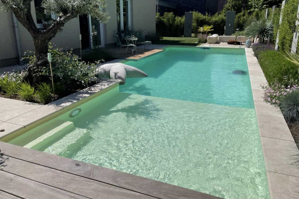 Ein rechteckiger Swimmingpool mit klarem Wasser, hellen Stufen und umliegenden Pflanzen und Grünflächen. In der Nähe des Hauses im Hintergrund sind ein Baum und ein kleiner Sitzbereich zu sehen, die das elegante Design der Pool-Folie unterstreichen.