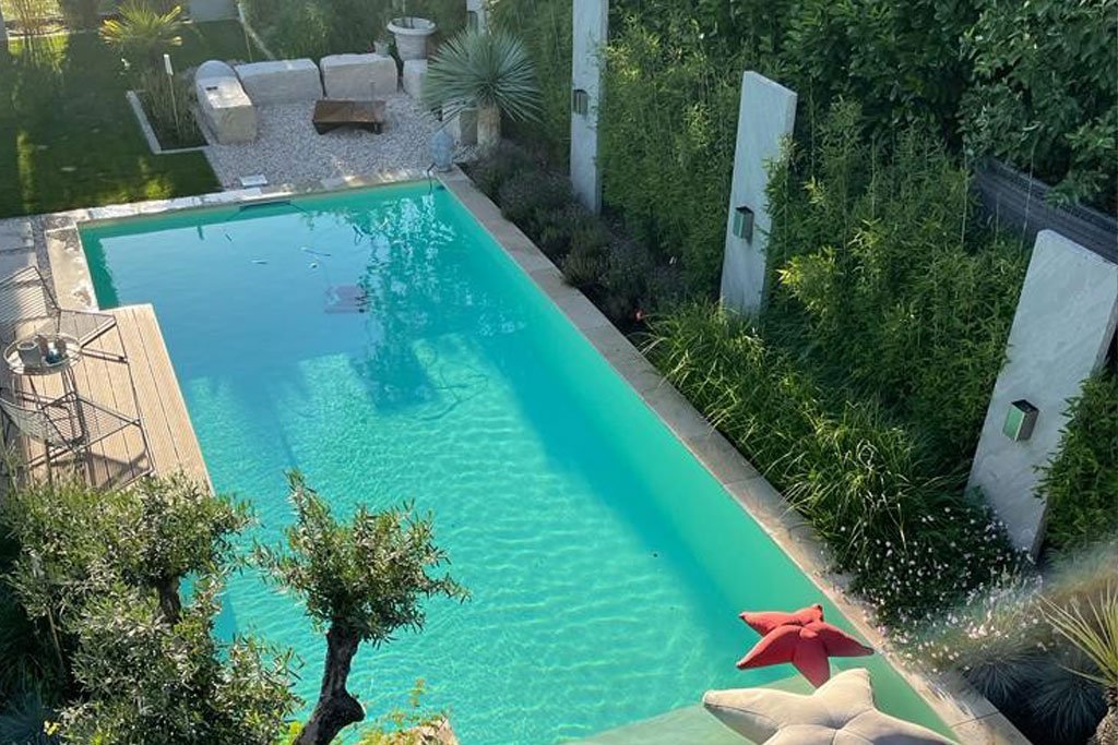 Ein rechteckiger Pool mit Folienauskleidung liegt in einem angelegten Garten, umgeben von Grünpflanzen und hohen Büschen. In der Nähe des Pools gibt es eine Sitzecke mit Kissen und einem roten sternförmigen Kissen sowie eine Steinbank in der Ecke.