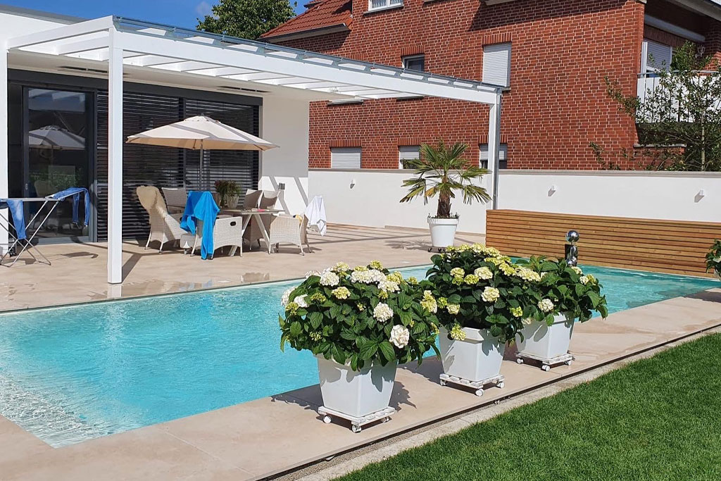 Ein moderner Hinterhof mit einem rechteckigen Pool, drei großen Topfpflanzen mit weißen Blumen, Liegestühlen unter einer Pergola und einem Backsteinhaus im Hintergrund.