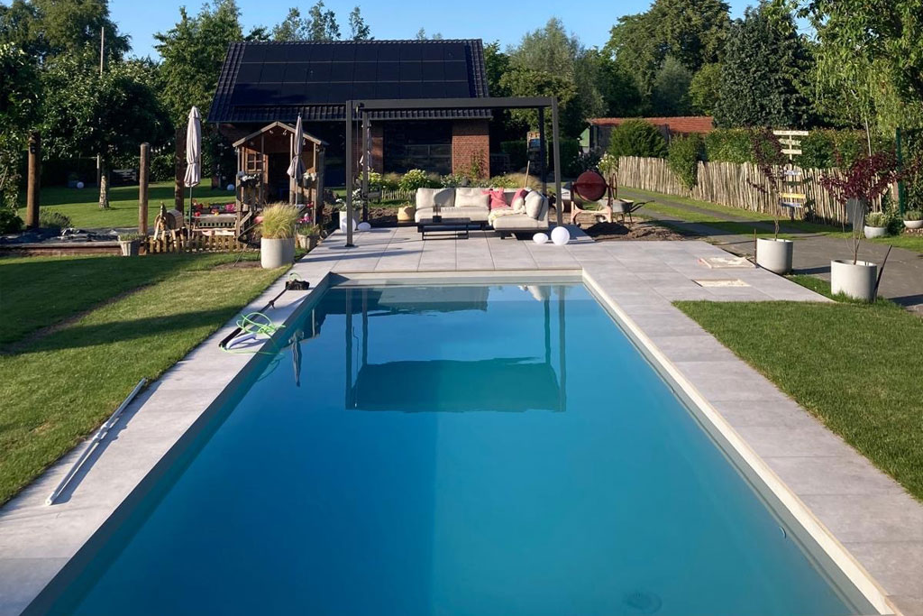 Ein rechteckiger Swimmingpool im Freien mit einer robusten Poolfolie ist von einer Steinterrasse und grünem Rasen umgeben. Poolreinigungsgeräte liegen an der Seite, während Terrassenmöbel, ein Grill und ein kleines Haus oder ein Schuppen zwischen Bäumen und Zäunen im Hintergrund stehen.
