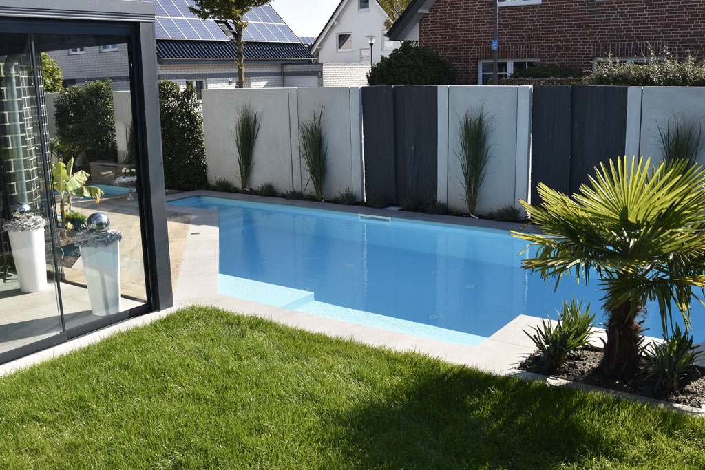 Ein moderner Hinterhof mit einem rechteckigen Pool, der von Gras, Pflanzen und hohen Sichtschutzwänden umgeben ist. Im Hintergrund sind ein Teil eines gläsernen Gebäudes und nahegelegene Häuser zu sehen, die eine elegante Pool-Oase bilden, die an einen stilvollen Teich erinnert.