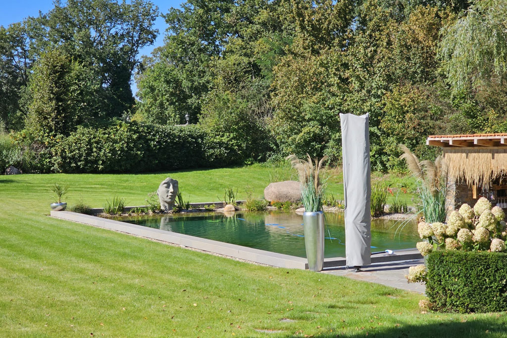 Ein rechteckiges, natürliches Schwimmbecken mit sauberen Rändern, oder Teich, ist von grünem Gras, Pflanzen und Bäumen umgeben. Eine große Kopfskulptur befindet sich in der Nähe des Wassers, und ein hohes, abgedecktes Objekt ruht am Beckenrand.