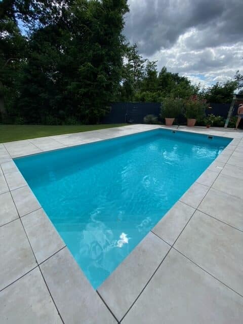 Ein rechteckiges Außenbecken mit klarem, blauem Wasser ist mit einer robusten PVC-Schwimmbadfolie Renolit Alkorplan 2000 (1,5 mm, acrylveredelt, gewebeverstärkt) ausgekleidet, umgeben von hellen Fliesen und Topfpflanzen unter einem bewölkten Himmel.